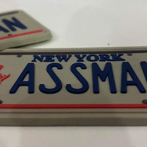 May include: A gray license plate with the text "NEW YORK ASSMAN" in blue letters. The plate has a red stripe and a small image of the Statue of Liberty in red.