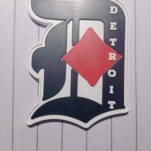 May include: A white and black 3D printed patch with a red diamond in the center. The patch has the word "DETROIT" written vertically on the right side.