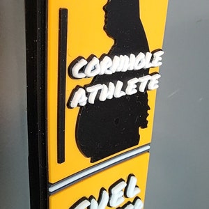 May include: A yellow and black sign with the text "Cornhole Athlete Fuel Center" in white letters. The sign features a silhouette of a person in a black cornhole pose.