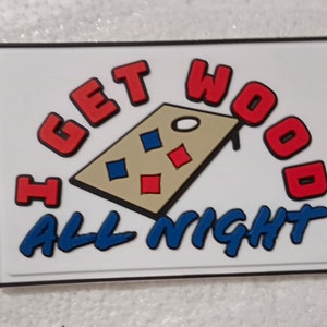 May include: A white rectangular sign with a black border. The sign has a red, blue, and tan illustration of a cornhole game board with the text "I GET WOOD ALL NIGHT" in red and blue.