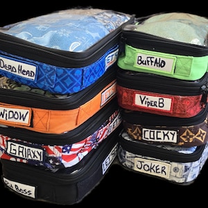 May include: A stack of six clear zippered pouches with different colorful fabric exteriors. Each pouch has a label with a different name written on it: Dead Head, Widow, Griaxy, Cocky, Joker, and Buffalo.