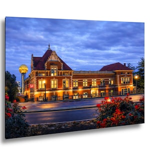 Puede incluir: Fotografía de un edificio de ladrillo con tejado de tejas rojas, iluminado por luces interiores, sobre un cielo crepuscular. Una estructura dorada en forma de esfera es visible en el fondo. Flores rojas bordean la carretera.