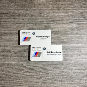 May include: Two white name tags with blue and white BMW logos. The top tag reads "BMW Car Club of America Boston Chapter" and "Michael Mangini, Director". The bottom tag reads "BMW Car Club of America Boston Chapter" and "Matt Bagedonow, Membership Chair".
