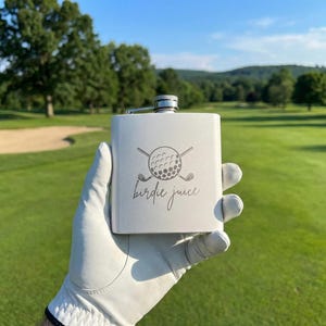 May include: A matte white metal flask featuring a laser-engraved graphic of a golf ball with two crossed golf clubs above the cursive text "birdie juice". The flask has a silver-colored screw-top cap and a rectangular body shape.