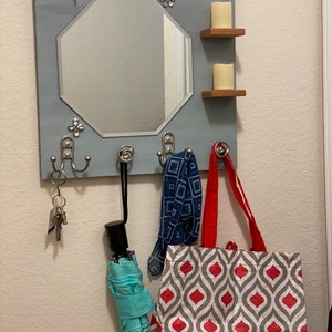 May include: A gray wooden wall mounted organizer with a mirror, two shelves, and three hooks. The mirror is octagonal and has a decorative floral design. The shelves are made of wood and have a light brown finish. The hooks are made of metal and have a silver finish. A blue and white patterned scarf, a red and gray patterned tote bag, and a light blue umbrella are hanging on the hooks. A set of keys is hanging on the leftmost hook.