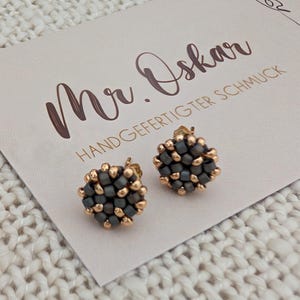 Könnte beinhalten: Ein Paar Ohrstecker bestehend aus einer kreisförmigen Anordnung kleiner, dunkelgrauer und goldfarbener Perlen. Die Ohrringe liegen auf einer hellen Karte mit dem Text Mr. Oskar HANDGEFERTIGTER SCHMUCK und einer Strichzeichnung eines Hundekopfes.