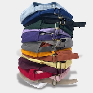 May include: A stack of 12 baseball caps in various colors, including blue, green, purple, gray, yellow, orange, maroon, red, pink, beige, and white. Each cap has a buckle closure.
