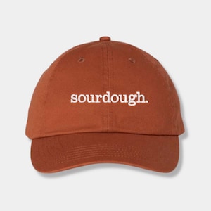 May include: A rust-colored baseball cap with the word "sourdough" embroidered in white across the front. The cap has a curved brim and a rounded crown, with a small button on top.
