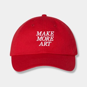 May include: A red baseball cap with the text "MAKE MORE ART" embroidered in white.