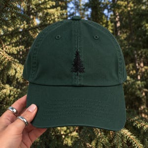 May include: A dark forest green baseball cap featuring a small, black embroidered pine tree design centered on the front panel. The cap has a structured crown, a curved brim with visible stitching, and a button at the top center.