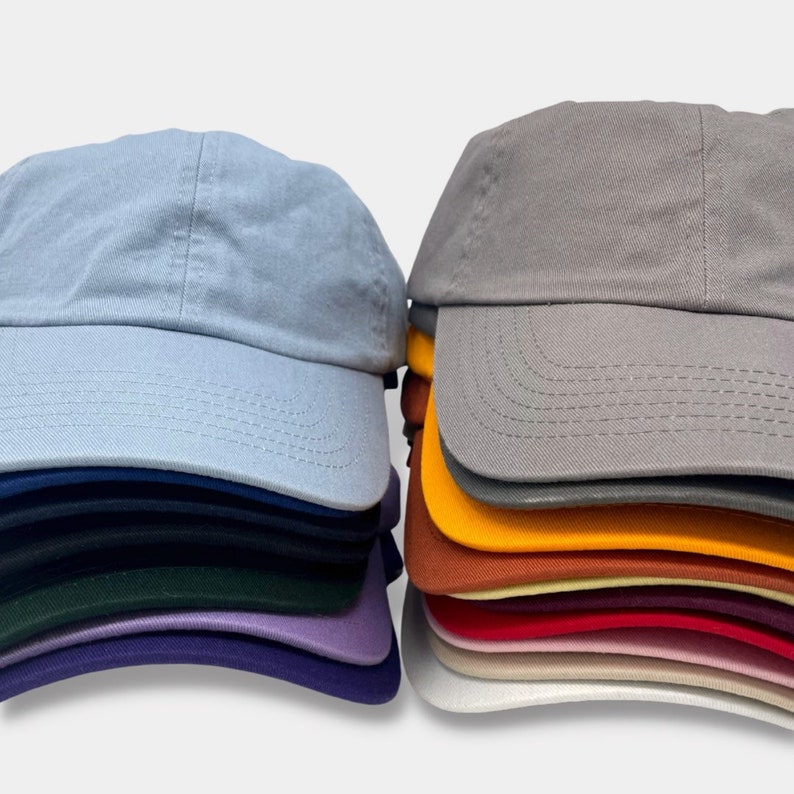 May include: A stack of blank, unbranded baseball caps in a variety of colors, including light blue, gray, yellow, orange, red, pink, white, and purple. The caps are made of a soft, cotton fabric and have a classic, six-panel design.