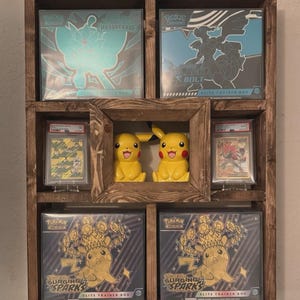 May include: A dark brown wooden display shelf containing four Pokemon Elite Trainer Boxes, two graded trading cards in protective cases, and two yellow Pikachu figurines. The boxes feature artwork from Twilight Masquerade, Black Bolt, and Surging Sparks.