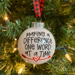 May include: White Christmas ornament with glitter and red accents. It reads "Making a difference one word at a time" with a red heart and arrows.