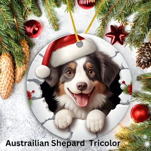 May include: A Christmas ornament featuring a tri-colored Australian Shepherd puppy wearing a Santa hat, peeking out of a cracked white circle. The ornament is surrounded by pine branches and red ornaments.