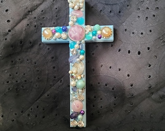 May include: A light blue cross adorned with various embellishments. The cross features pearls, iridescent beads, and gemstones in shades of pink, blue, and gold. The decorative cross is a religious item.