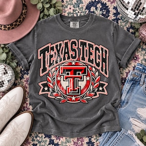 Könnte beinhalten: Ein anthrazitfarbenes Kurzarm-T-Shirt mit dem Text Texas Tech in einer fetten, schwarz-roten, gebogenen Schrift. Die Grafik in der Mitte zeigt ein rot-schwarzes Doppel-T-Logo, umgeben von einem kreisförmigen Lorbeerkranz mit kleinen weißen Sternen.
