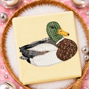 May include: A light yellow fabric square featuring a mallard duck appliqué. The duck has a green gingham head, a yellow polka-dot beak, a brown floral chest, and wings made of white striped, black lace, and dark grey patterned fabric patches.