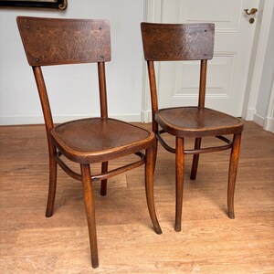 May include: A pair of brown wooden chairs featuring curved backrests with intricate embossed floral patterns and circular seats with subtle diagonal line detailing. Each chair is supported by four slender, slightly flared legs connected by curved supports.