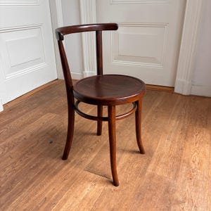 Peut inclure: Une chaise en bois brun foncé dotée d'une assise circulaire et d'un dossier incurvé soutenu par un montant vertical unique. La chaise possède quatre pieds fins légèrement courbés vers l'extérieur, reliés par un anneau de support.