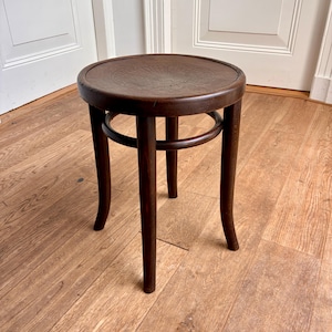 May include: A dark brown wooden stool featuring a circular seat with a decorative embossed pattern in the center. The stool is supported by four curved legs connected by a circular wooden ring brace positioned beneath the seat for stability.
