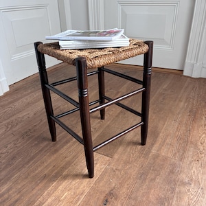 Op de afbeelding: Een vierkante kruk met een donkerbruin houten frame met vier poten en horizontale steunbalken. De zitting is gemaakt van gevlochten natuurlijk vezelmateriaal. Een stapel van drie witte tijdschriften ligt plat bovenop de zitting van de kruk.