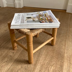 Op de afbeelding: Een kleine houten kruk met een geweven zitting ondersteunt een stapel "The World of Interiors" tijdschriften. De tijdschriften zijn wit met zwarte tekst. De kruk is gemaakt van lichtgekleurd hout.
