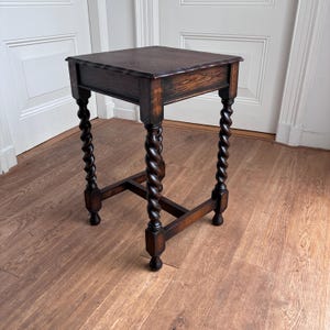 Op de afbeelding: Een vierkante houten bijzettafel met een donkerbruine afwerking en zichtbare houtnerf. De tafel heeft vier poten met een opvallend gedraaid ontwerp, verbonden door een onderste H-vormige dwarsbalk, en rust op kleine, ronde voetjes aan de basis.