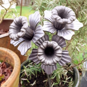 May include: A metal sculpture of three stylised flowers with grey petals and black centres. The flowers are arranged on a stem and are set against a background of green foliage.
