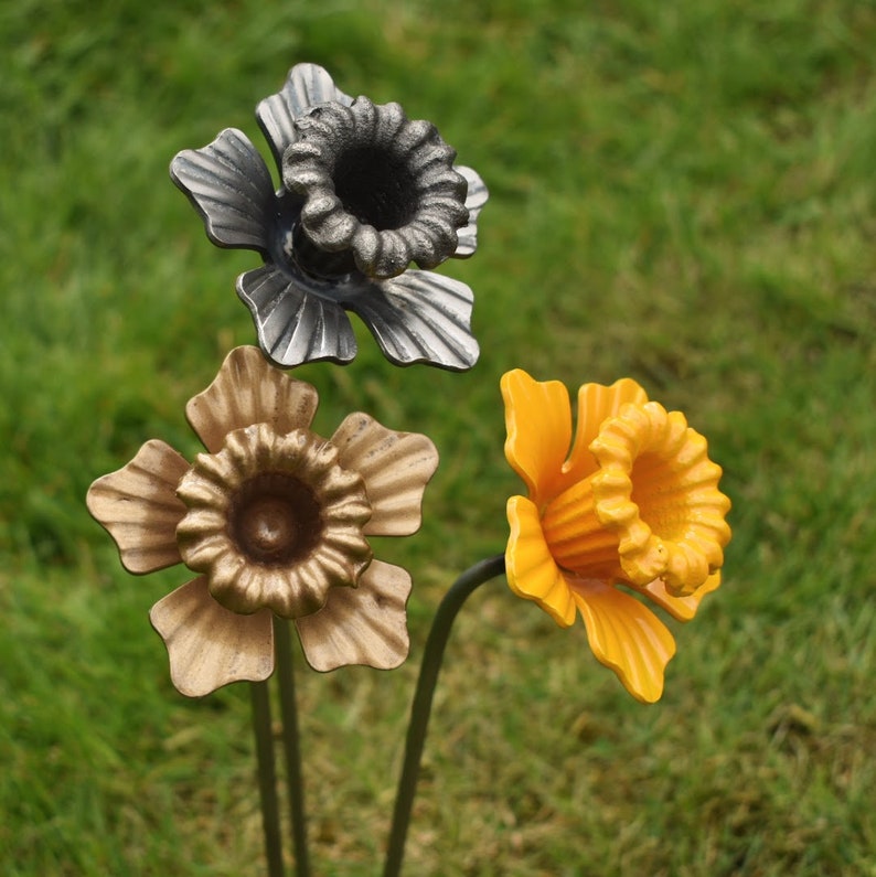 Puede incluir: Tres flores de metal con tallos. Las flores tienen forma de narcisos. Una flor es plateada, otra es dorada y otra es amarilla.