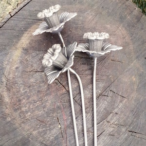 Puede incluir: Tres flores de metal plateado con tallos largos. Las flores tienen un dise&ntilde;o delicado y detallado y est&aacute;n dispuestas sobre una superficie de madera.