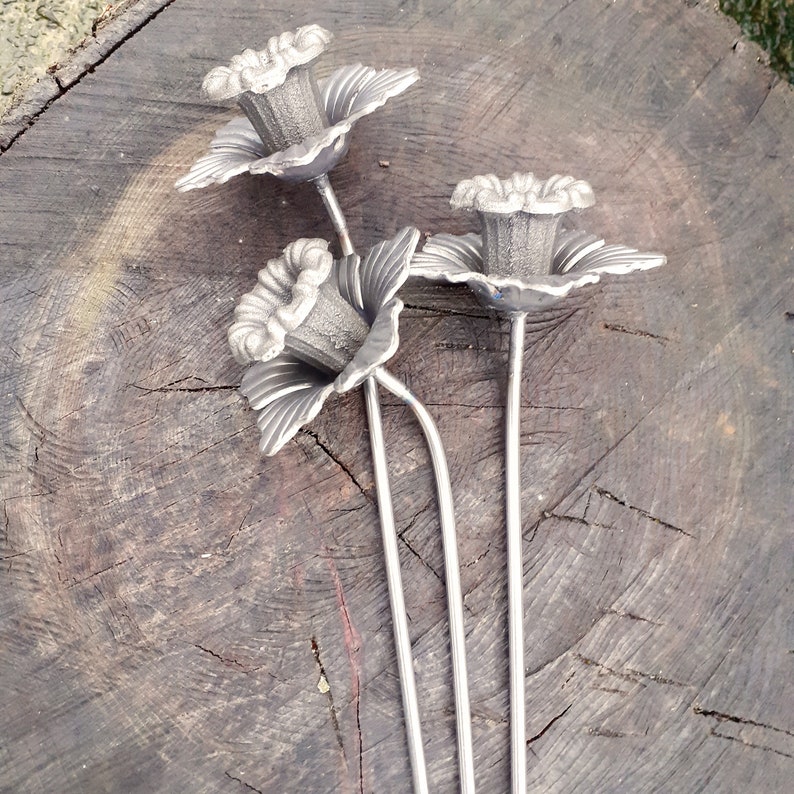 Puede incluir: Tres flores de metal plateado con tallos largos. Las flores tienen un dise&ntilde;o delicado y detallado y est&aacute;n dispuestas sobre una superficie de madera.