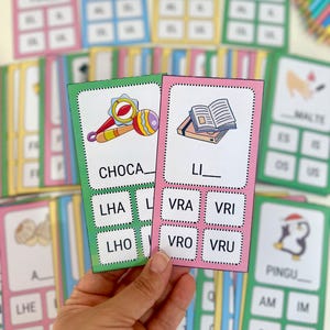 Pode incluir: Dois cartões de memória rosa e verde com as palavras "CHOCA" e "LI" escritas neles.  Abaixo das palavras, há quatro caixas com as letras "LHA", "LHO", "VRA", "VRI", "VRO" e "VRU" escritas nelas.  Os cartões de memória estão sendo segurados por uma mão.