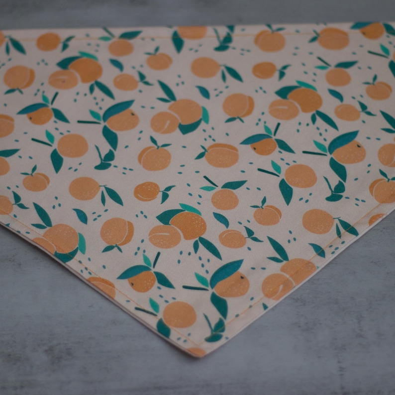 May include: A peach-colored bandana with a repeating pattern of orange peaches and green leaves. The bandana is made of a soft, lightweight fabric and has a triangular shape.