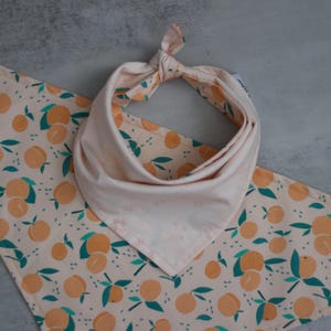 May include: A peach-colored bandana with a peach and leaf pattern. The bandana is tied in a knot at the top and has a white background.
