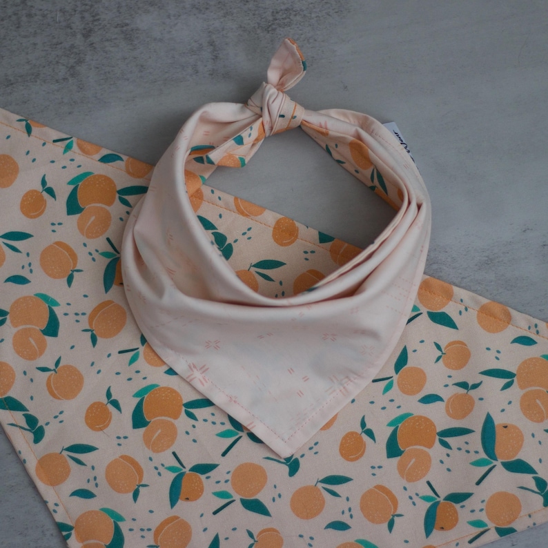 May include: A peach-colored bandana with a peach and leaf pattern. The bandana is tied in a knot at the top and has a white background.