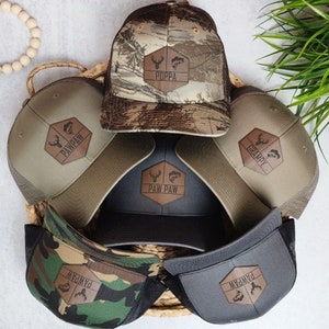 May include: A collection of baseball caps with mesh backs and leather patches. The patches feature text like "Poppa," "Pawpaw," and "Grampy," along with deer and fish graphics. The caps are in various colors, including camouflage, gray, and tan.