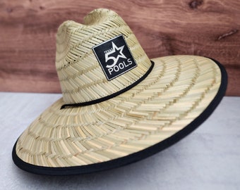May include: A light tan straw hat with a wide brim and a black fabric trim along the edge. The hat features a black square patch on the front with a white star graphic and the text Texas 5 Pools printed in white lettering on the front crown.