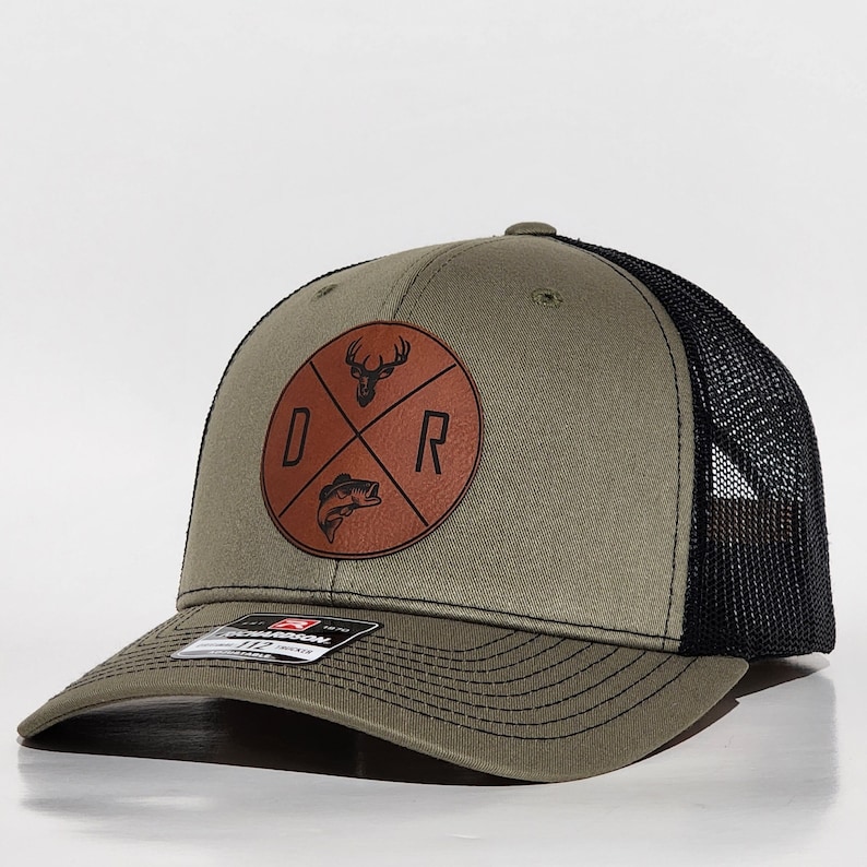 May include: A green and black trucker hat with a brown leather patch featuring a deer, fish, and the letters "D" and "R".