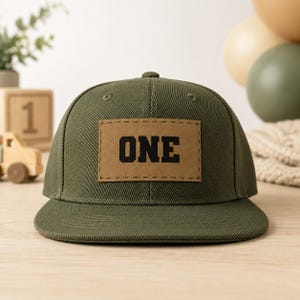 May include: An olive green baseball cap featuring a rectangular tan patch centered on the front panel. The patch has a stitched border and displays the word ONE in bold black capital letters. The cap has a structured crown and a flat brim.