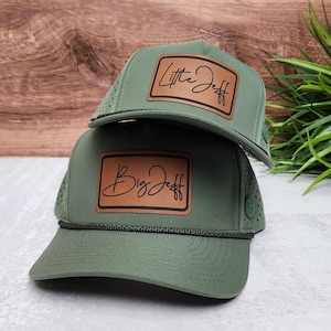 May include: Two olive green baseball caps with perforated side panels and a dark green rope detail across the brim. Each cap features a rectangular tan leather patch with black text, one reading Little Jeff and the other reading Big Jeff.