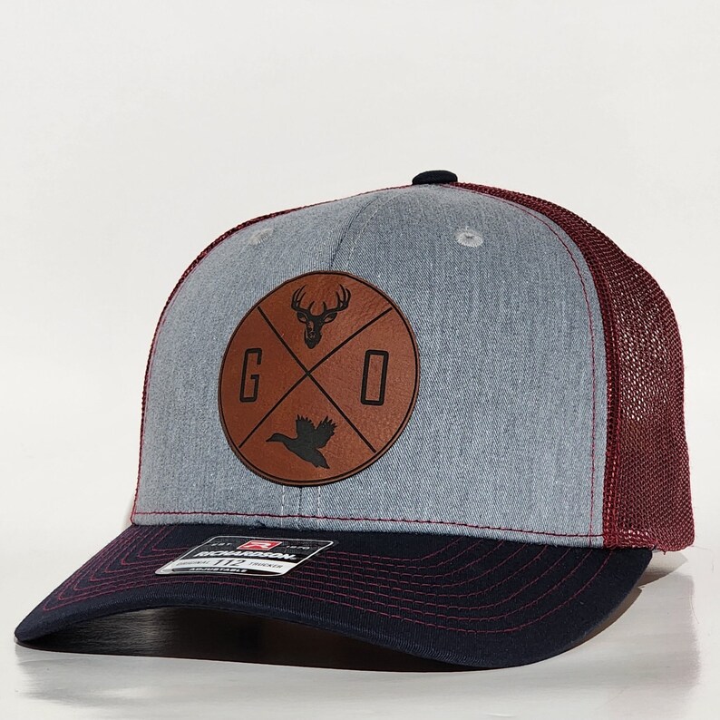 May include: A gray and red trucker hat with a brown leather patch featuring a deer head, a bird, and the letters "GO".
