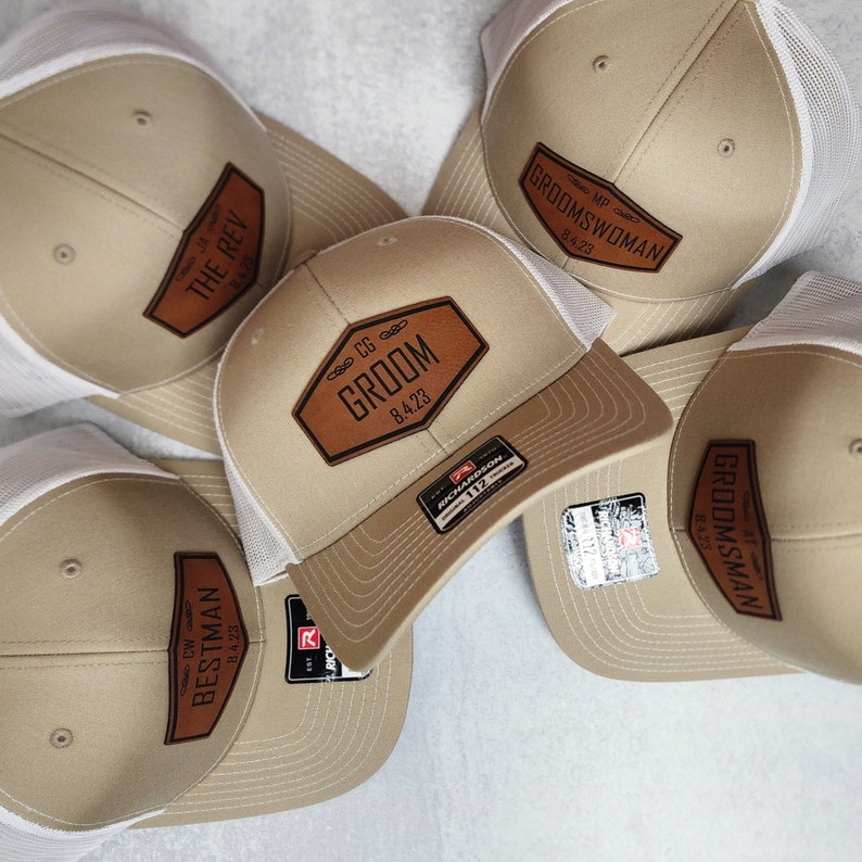 May include: Tan baseball caps with white mesh backs and leather patches. The patches feature text such as "GROOM," "BESTMAN," and "GROOMSMAN," along with dates. The caps have a curved brim and a Richardson 112 tag.