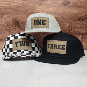 May include: Three baseball caps with brown patches that say "ONE", "TWO", and "THREE" in black letters. The caps are gray, black and white checkered, and black.