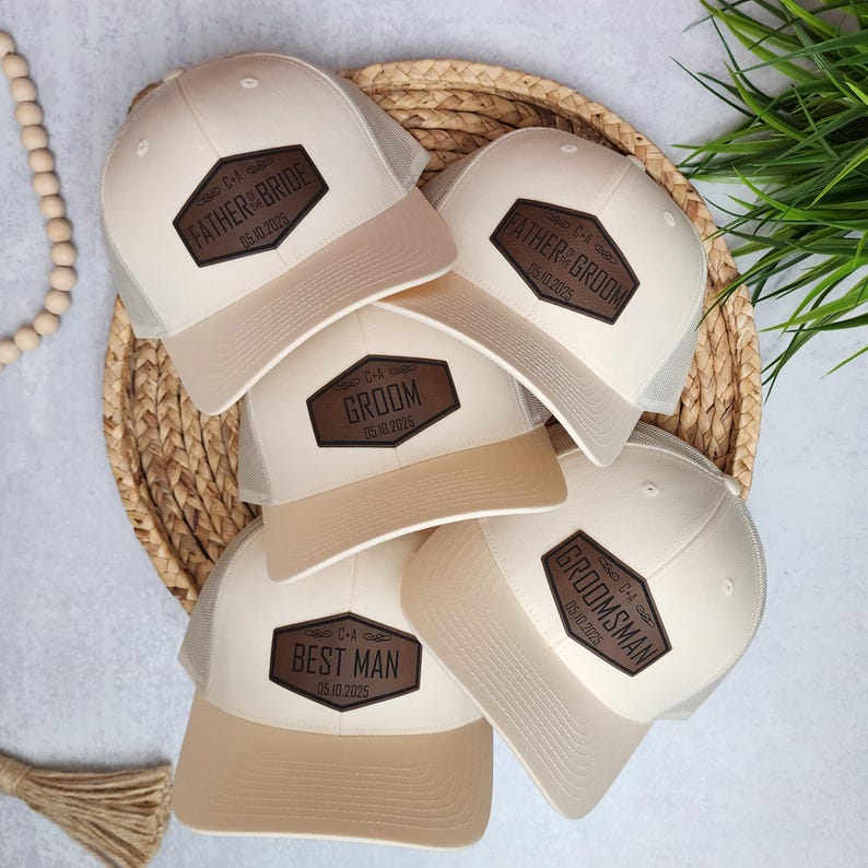 May include: A collection of beige and white trucker hats with mesh backs and brown leather patches. The patches display text such as "Father of the Bride," "Groom," "Best Man," and "Groomsman," along with a date. The hats are arranged on a woven basket.