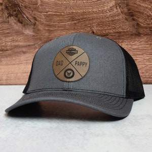 May include: A gray and black trucker hat with a brown leather patch that says "DAD" and "PAPPY" with a Harley Davidson logo in the center.