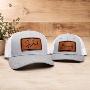 May include: Two gray and white trucker hats featuring brown rectangular patches. The larger hat has the word Dad in black script on the patch, while the smaller hat features the name Bennett in black script on its matching brown patch.