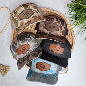 May include: Five camouflage trucker hats with a brown leather patch that says "DAD" on each hat. The hats are arranged in a circle on a woven straw surface.