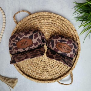 May include: Two camouflage trucker hats with brown leather patches. The larger hat has the word "DAD" on the patch, and the smaller hat has the name "WAYLON" on the patch.
