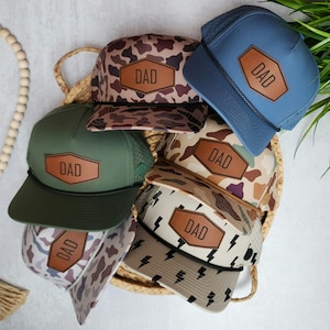 May include: Five different baseball caps with a camouflage pattern and a leather patch that says "DAD". The caps are arranged in a circle on a woven basket.