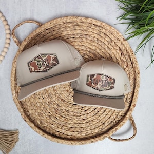 May include: Two beige baseball caps with a camouflage design and the names "DAD" and "WAYLON" printed on them. The caps have a brown brim and a decorative rope detail. They are displayed on a woven tray.